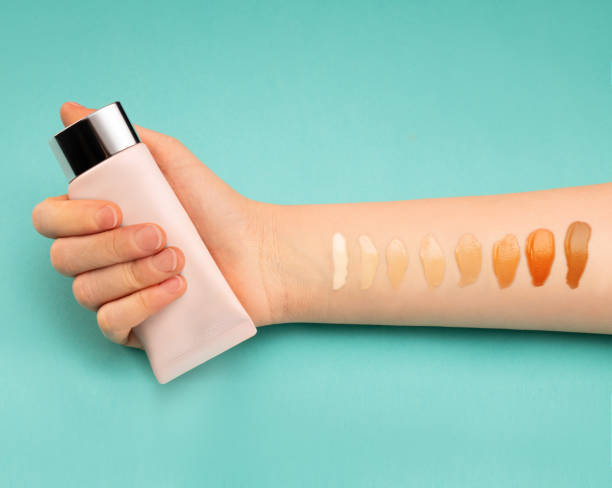 Person holding foundation bottle with swatches of different shades on their arm against a teal background.